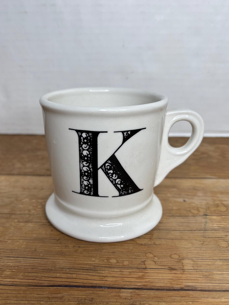Pu&ograve; includere: Tazza in ceramica bianca con una lettera "K" nera. La tazza ha una base arrotondata e un manico. La lettera "K" &egrave; decorata con motivi decorativi. La tazza &egrave; su una superficie di legno.