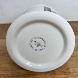 Pu&ograve; includere: Base di una tazza in ceramica bianca con un logo circolare raffigurante un'illustrazione di un uccello. Il logo &egrave; circondato dal testo "MICROWAVE & DISHWASHER SAFE" e "FABRIQUE EN CHINE". La tazza &egrave; su una superficie di legno.