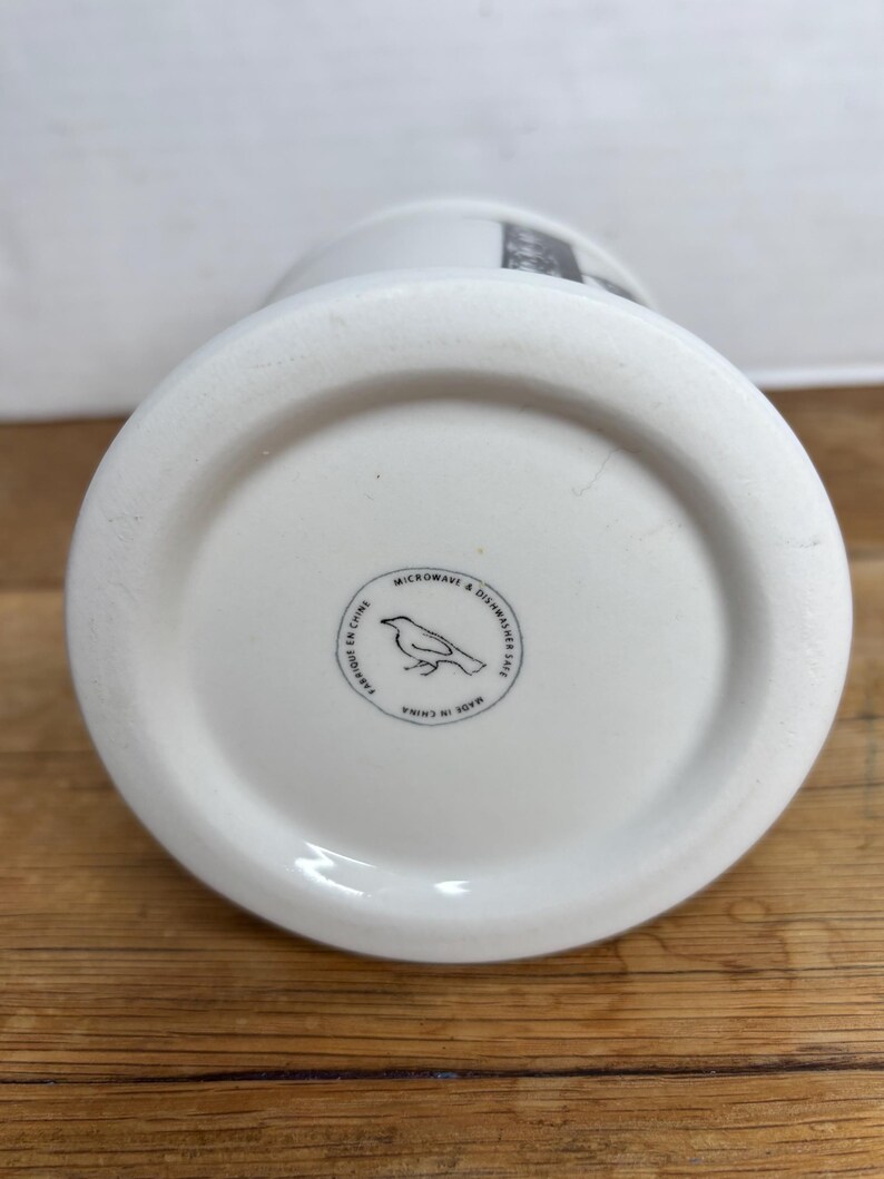 Pu&ograve; includere: Base di una tazza in ceramica bianca con un logo circolare raffigurante un'illustrazione di un uccello. Il logo &egrave; circondato dal testo "MICROWAVE & DISHWASHER SAFE" e "FABRIQUE EN CHINE". La tazza &egrave; su una superficie di legno.