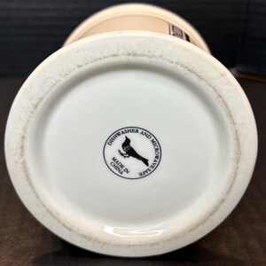 K&ouml;nnte beinhalten: Unterseite einer wei&szlig;en Keramik-Tasse mit beigem Rand. Der Boden hat ein schwarz-wei&szlig;es kreisf&ouml;rmiges Logo mit einem Vogel und dem Text "Dishwasher and Microwave Safe" und "Made in China."