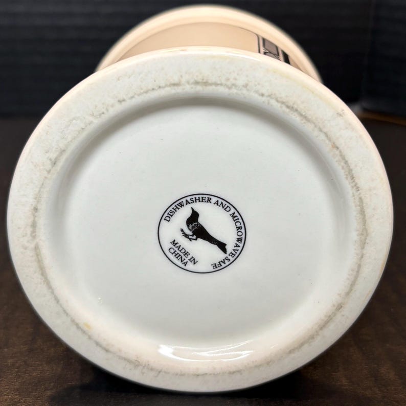 K&ouml;nnte beinhalten: Unterseite einer wei&szlig;en Keramik-Tasse mit beigem Rand. Der Boden hat ein schwarz-wei&szlig;es kreisf&ouml;rmiges Logo mit einem Vogel und dem Text "Dishwasher and Microwave Safe" und "Made in China."