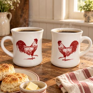 May include: Two off-white ceramic mugs filled with dark coffee, each featuring a detailed red illustration of a rooster. The mugs have a curved handle and a wide base, sitting on a wooden surface alongside a plate of biscuits and butter.