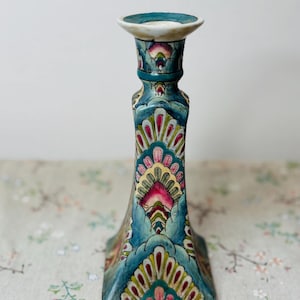 May include: A tall, ornate candle holder with a square base and a flared top. The ceramic piece features a teal background with a floral pattern in pink, yellow, and green. The top is a light teal color.