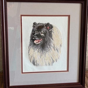 May include: Framed cross-stitch portrait of a Keeshond dog. The dog has a black and gray coat with a light tan ruff. The dog's tongue is visible. The artwork is framed in a dark brown wooden frame with a gray mat.