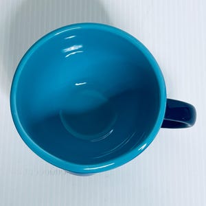 K&ouml;nnte beinhalten: Eine Draufsicht auf eine Keramiktasse mit einem leuchtend t&uuml;rkisfarbenen Inneren und einer glatten, gl&auml;nzenden Oberfl&auml;che. Die Tasse hat eine abgerundete Schalenform und einen dunkelblauen Henkel an der Seite, was ein schlichtes Design zeigt.