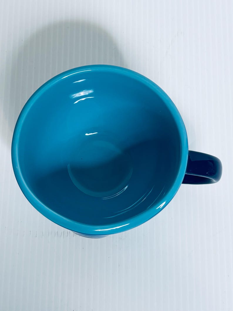 K&ouml;nnte beinhalten: Eine Draufsicht auf eine Keramiktasse mit einem leuchtend t&uuml;rkisfarbenen Inneren und einer glatten, gl&auml;nzenden Oberfl&auml;che. Die Tasse hat eine abgerundete Schalenform und einen dunkelblauen Henkel an der Seite, was ein schlichtes Design zeigt.