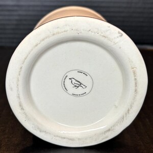 May include: A cream-colored ceramic vase with a wide base and a narrower top. The bottom of the vase features a circular emblem with a bird illustration and the words "Hand Wash", "Fabriqu&eacute; en Chine", and "Made in China".