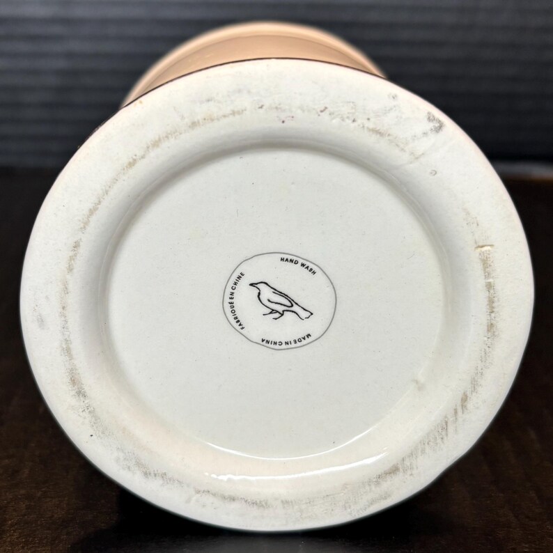 May include: A cream-colored ceramic vase with a wide base and a narrower top. The bottom of the vase features a circular emblem with a bird illustration and the words "Hand Wash", "Fabriqu&eacute; en Chine", and "Made in China".