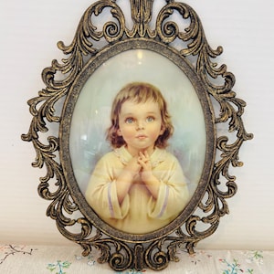 May include: An antique oval-shaped picture frame with a detailed, ornate bronze finish. The frame surrounds a portrait of a young child with light brown hair, wearing a yellow dress, and clasping their hands. The background is a soft blue.