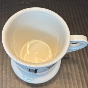 K&ouml;nnte beinhalten: Wei&szlig;er Keramikbecher mit Henkel und Untertasse. Der Becher hat ein schlichtes Design mit hellem Innenraum und einem dekorativen Muster auf der Untertasse. Der Becher ist f&uuml;r Getr&auml;nke konzipiert.