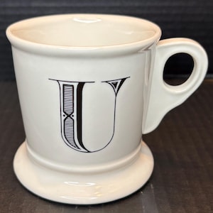 K&ouml;nnte beinhalten: Cremefarbene Keramiktasse mit dem schwarzen Buchstaben "U". Die Tasse hat einen abgerundeten Boden und einen gebogenen Henkel. Das "U" ist ein dekoratives Design. F&uuml;r Hei&szlig;getr&auml;nke geeignet.