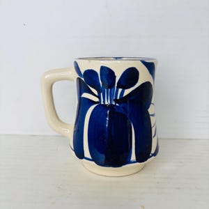 May include: Cream-colored ceramic mug with a large, stylized blue floral design. The mug has a rounded base, a straight body, and a sturdy handle. The blue design is painted in bold strokes, creating a striking contrast against the cream background.