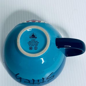 K&ouml;nnte beinhalten: Ein hellblauer Keramikbecher mit einem dunkelblauen Henkel. Der Boden zeigt das Disney-Logo, den Text MADE IN THAILAND, FABRIQU&Eacute; EN THAILANDE, HECHO EN TAILANDIA und den Code FAC-024367-20302. Das Wort Stitch ist in Schwarz auf der Seite gedruckt.