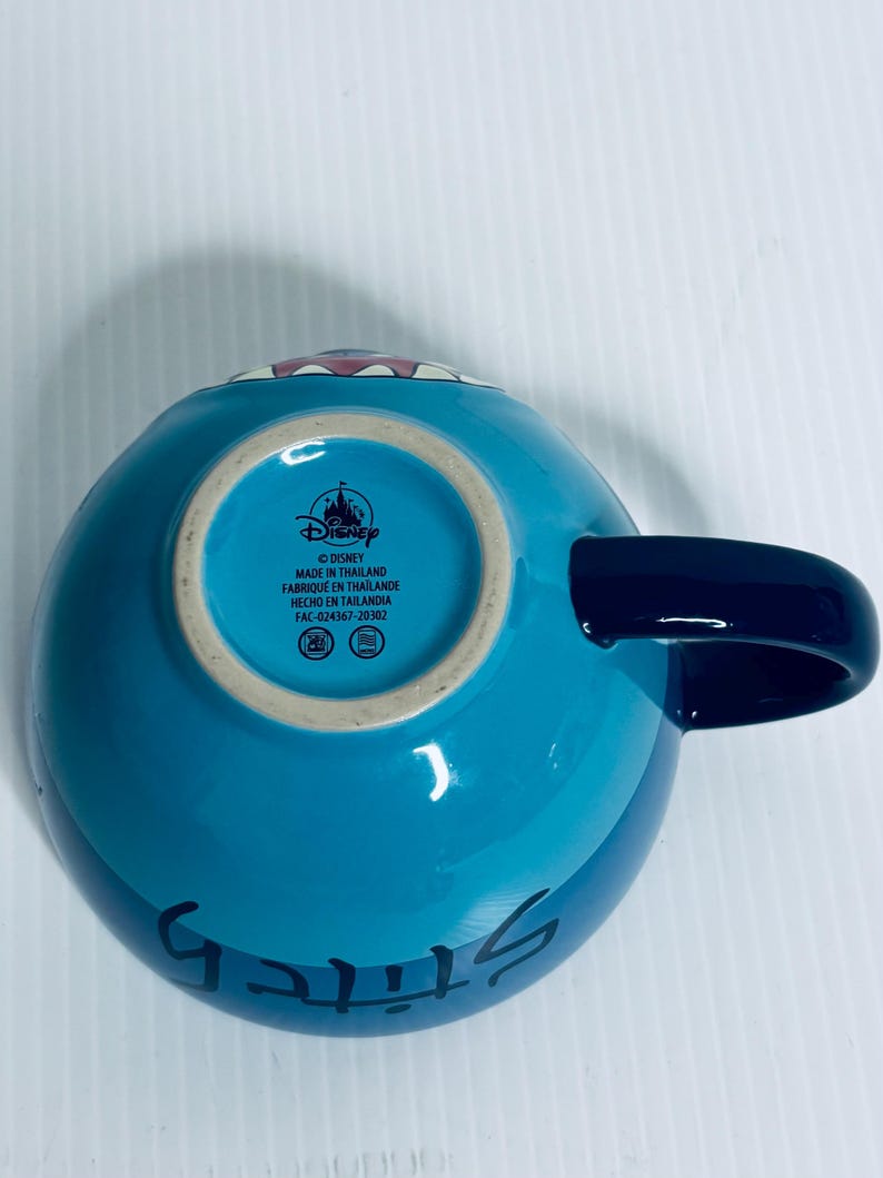 K&ouml;nnte beinhalten: Ein hellblauer Keramikbecher mit einem dunkelblauen Henkel. Der Boden zeigt das Disney-Logo, den Text MADE IN THAILAND, FABRIQU&Eacute; EN THAILANDE, HECHO EN TAILANDIA und den Code FAC-024367-20302. Das Wort Stitch ist in Schwarz auf der Seite gedruckt.