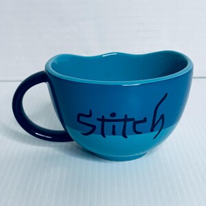 K&ouml;nnte beinhalten: Ein zweifarbiger blauer Keramikbecher mit gewelltem Rand und einem dunkelblauen Henkel. Auf der Au&szlig;enseite ist das Wort Stitch in einer dunkelblauen, handschriftlichen Schriftart in der Mitte zu sehen. Das Innere des Bechers ist einfarbig t&uuml;rkis.
