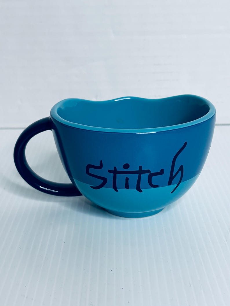 K&ouml;nnte beinhalten: Ein zweifarbiger blauer Keramikbecher mit gewelltem Rand und einem dunkelblauen Henkel. Auf der Au&szlig;enseite ist das Wort Stitch in einer dunkelblauen, handschriftlichen Schriftart in der Mitte zu sehen. Das Innere des Bechers ist einfarbig t&uuml;rkis.