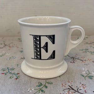 Puede incluir: Taza de cer&aacute;mica blanca con la letra "E" negra en una fuente decorativa. La taza tiene un asa redondeada y una base de pedestal. La taza est&aacute; sobre una superficie con estampado floral.