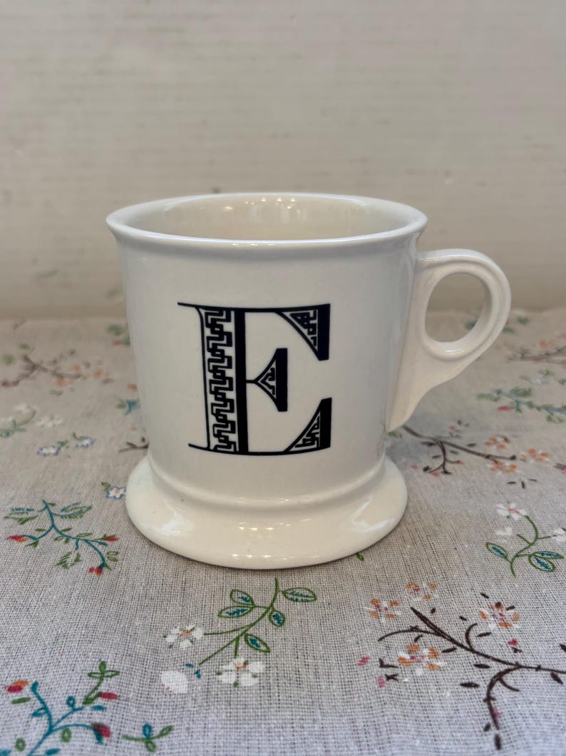 Puede incluir: Taza de cer&aacute;mica blanca con la letra "E" negra en una fuente decorativa. La taza tiene un asa redondeada y una base de pedestal. La taza est&aacute; sobre una superficie con estampado floral.