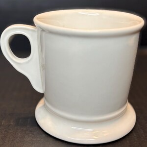 K&ouml;nnte beinhalten: Eine wei&szlig;e Keramiktasse mit rundem Boden und Henkel. Die Tasse hat ein schlichtes, klassisches Design mit glatter, gl&auml;nzender Oberfl&auml;che. Der Henkel ist an der Seite der Tasse befestigt und hat eine runde &Ouml;ffnung.