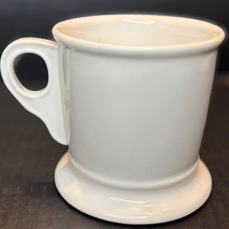 K&ouml;nnte beinhalten: Eine wei&szlig;e Keramiktasse mit rundem Boden und Henkel. Die Tasse hat ein schlichtes, klassisches Design mit glatter, gl&auml;nzender Oberfl&auml;che. Der Henkel ist an der Seite der Tasse befestigt und hat eine runde &Ouml;ffnung.