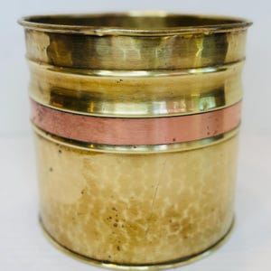 May include: A cylindrical brass planter with a hammered texture. The planter features a copper band around the middle and a smooth brass rim. The overall design is a mix of textures and colors, suitable for home decor.