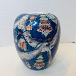 May include: A blue ceramic vase with a decorative pattern. The vase features a dark blue background with a design of white ribbons and colorful, ornate shapes. The vase has a rounded shape and a white interior.