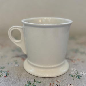 Puede incluir: Una taza de cer&aacute;mica blanca con un asa redondeada y una base de pedestal. La taza tiene un dise&ntilde;o simple y cl&aacute;sico y un acabado liso y brillante. La taza probablemente se usa para beber caf&eacute; o t&eacute;.