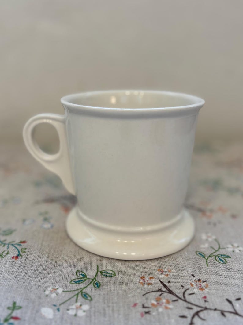 Puede incluir: Una taza de cer&aacute;mica blanca con un asa redondeada y una base de pedestal. La taza tiene un dise&ntilde;o simple y cl&aacute;sico y un acabado liso y brillante. La taza probablemente se usa para beber caf&eacute; o t&eacute;.