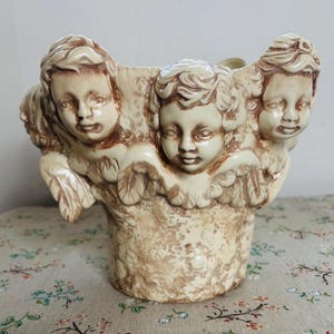 May include: A beige ceramic planter featuring three cherubic faces with detailed wings. The planter has a textured, aged appearance, with brown accents highlighting the cherubs' features. The container is a decorative item.