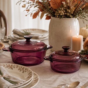 May include: Two round, deep purple glass casserole dishes with matching lids. Each dish features two side handles and a textured circular pattern on the center of the lid. The glass is translucent, allowing light to pass through the deep violet hue.