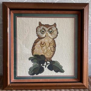 May include: Framed cross-stitch artwork featuring a detailed owl perched on a branch with green leaves. The owl has brown and tan feathers, large yellow eyes, and is set against a neutral background. The frame is a warm brown with a green and tan matting.