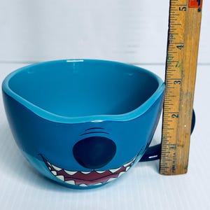 K&ouml;nnte beinhalten: Ein leuchtend blauer Keramikbecher in Form der Figur Stitch, der ein breites Grinsen mit sichtbaren Z&auml;hnen und eine schwarze Nase aufweist. Ein vertikal neben dem Becher platziertes Holzlineal zeigt eine H&ouml;he von etwa 8,9 cm an.