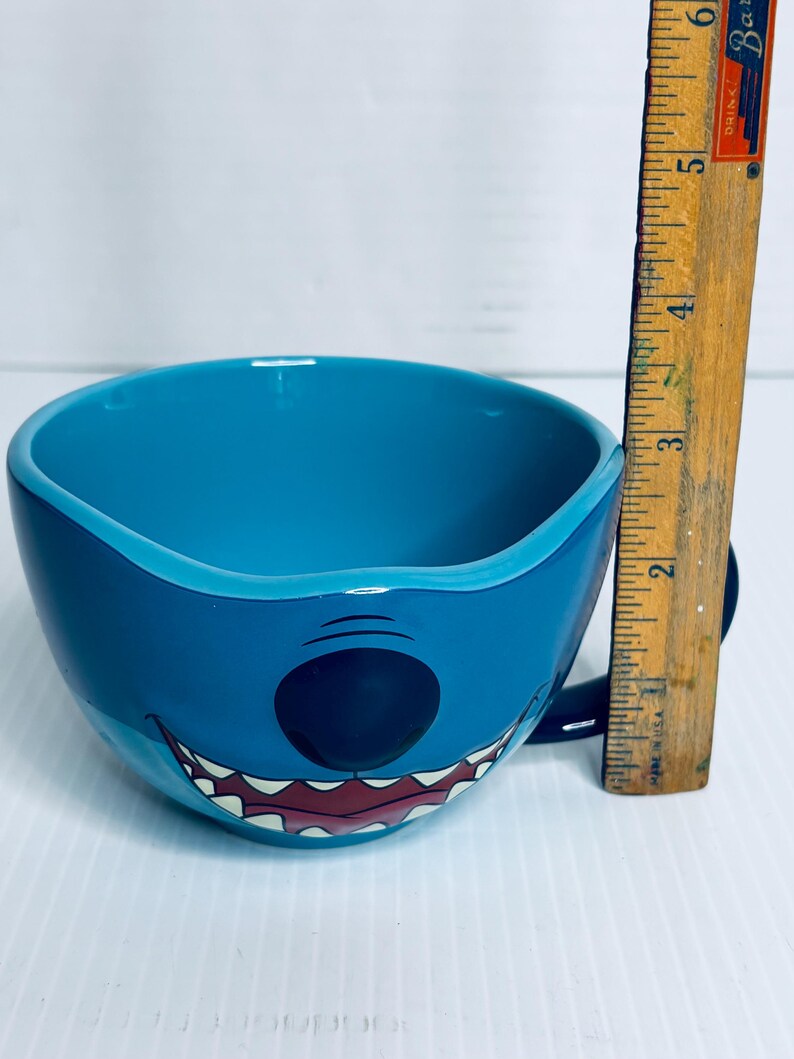 K&ouml;nnte beinhalten: Ein leuchtend blauer Keramikbecher in Form der Figur Stitch, der ein breites Grinsen mit sichtbaren Z&auml;hnen und eine schwarze Nase aufweist. Ein vertikal neben dem Becher platziertes Holzlineal zeigt eine H&ouml;he von etwa 8,9 cm an.