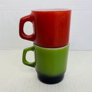 May include: Two vintage stackable coffee mugs. The top mug is red, and the bottom mug is green with a black base. Both mugs have a handle and a white rim. The mugs are stacked on top of each other.