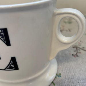Puede incluir: Taza de cer&aacute;mica blanca con base redondeada y asa circular. La taza presenta letras negras en el lateral. El asa est&aacute; fijada al lateral de la taza. La taza est&aacute; sobre una superficie con estampado floral.