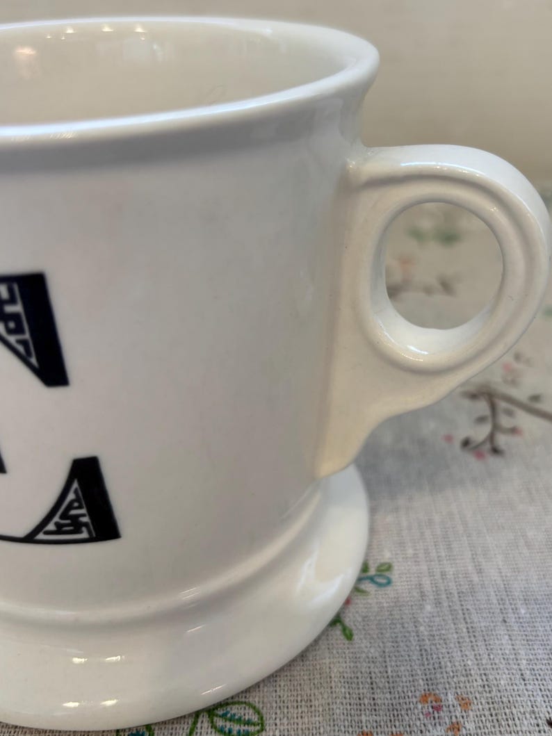 Puede incluir: Taza de cer&aacute;mica blanca con base redondeada y asa circular. La taza presenta letras negras en el lateral. El asa est&aacute; fijada al lateral de la taza. La taza est&aacute; sobre una superficie con estampado floral.
