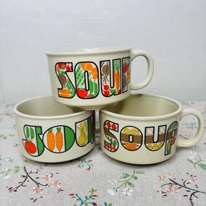 May include: Three cream-colored soup bowls with handles, each displaying the word "SOUP" in large, colorful letters. The letters are filled with illustrations of vegetables like carrots, tomatoes, and onions. The bowls are stacked on a floral patterned surface.