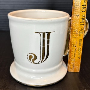 May include: A white ceramic mug with a gold handle and trim, featuring a large gold letter "J". The mug has a pedestal base and a ruler is next to it. The ruler has the text "A GOOD RULE Do Unto Other".