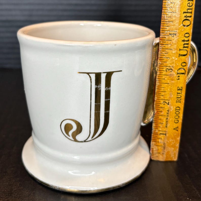 May include: A white ceramic mug with a gold handle and trim, featuring a large gold letter "J". The mug has a pedestal base and a ruler is next to it. The ruler has the text "A GOOD RULE Do Unto Other".
