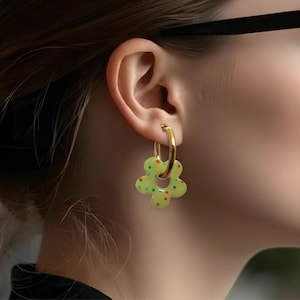 Peut inclure: Boucles d'oreilles créoles dorées avec un charm en forme de fleur. Le charm est vert clair avec des pois rouges et jaunes. Un accessoire élégant.