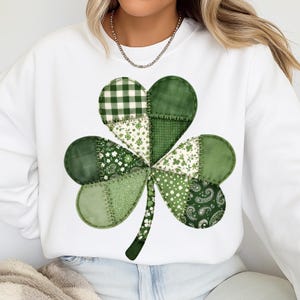 Peut inclure: Sweat-shirt blanc orné d'un grand trèfle à quatre feuilles en patchwork. Le trèfle est composé de différents tissus verts, dont des motifs vichy, floraux et cachemire, cousus ensemble. Le motif est centré sur le devant du sweat-shirt.