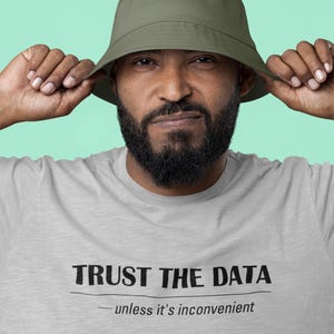 Könnte beinhalten: Ein hellgraues T-Shirt mit dem Text "TRUST THE DATA" und dem Slogan "unless it's inconvenient". Die Person auf dem Bild trägt einen grünen Fischerhut.