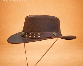 Puede incluir: Sombrero de cuero marrón con ala ancha y banda de cuero. La banda está adornada con pequeños tachuelas redondas de color latón y detalles metálicos decorativos. El sombrero tiene una correa para la barbilla y el texto "KhoobArts".