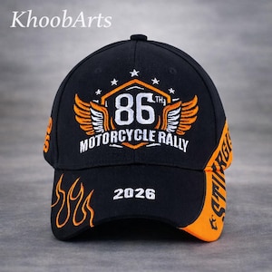 May include: A black baseball cap featuring orange and white embroidery. The front displays an 86th Motorcycle Rally emblem with wings, stars, and the year 2026. Orange flame patterns decorate the brim, and orange side panels feature text.