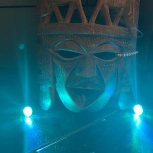 May include: A carved wooden mask with a stylized face and two carved animal figures at the top. The mask is brown with a beaded accent and is illuminated with blue light.