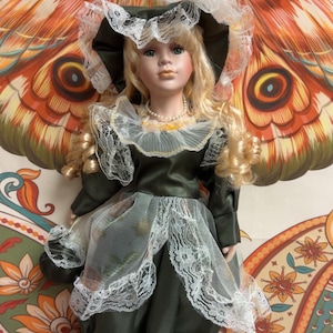 May include: A porcelain doll with blonde curly hair, wearing a dark green satin dress with white lace trim, a matching wide-brimmed hat with white feathers, and a pearl necklace. The doll stands on a white circular base and holds a small dark bag.