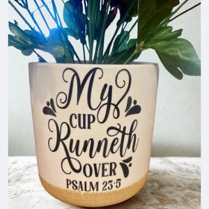 May include: A cream-coloured ceramic flower pot with the words "My Cup Runneth Over" and "Psalm 23:5" in black script. The base of the pot is tan. The pot is filled with green and blue artificial flowers.