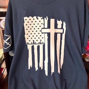 Pode incluir: Camiseta azul marinho com um design de bandeira americana desgastada e uma cruz branca. A bandeira inclui estrelas e listras brancas. Um pequeno emblema com espadas cruzadas está na manga esquerda.