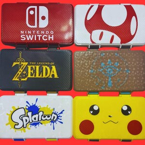 May include: Six rectangular game cartridge cases arranged in two columns. The top left is red with the Nintendo Switch logo, top right is white with a red mushroom, middle left is black with The Legend of Zelda logo, middle right is brown with a blue eye symbol, bottom left is white with the Splatoon logo, and bottom right is yellow with a Pikachu face.