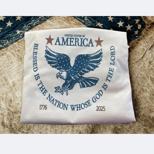 以下が含まれることがあります： 青いワシのデザインが施された白いTシャツ。「UNITED STATES OF AMERICA」と「BLESSED IS THE NATION WHOSE GOD IS THE LORD」の文字入り。Tシャツには1776年と2025年の日付も表示されています。